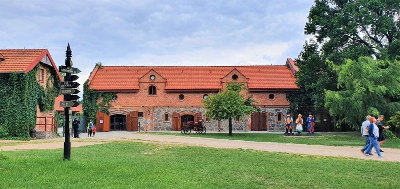 Widok na stajnie, budynek z czerwonej cegły i czerwoną dachówką, przed budynkiem stoją posągi ludzi, wokół spacerujący turyści - Muzeum Wsi Mazowieckiej w Sierpcu