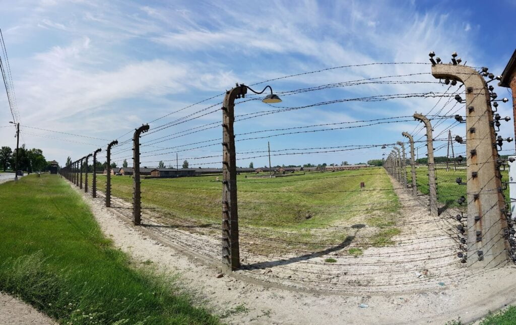 Auschwitz-Birkenau - Ogrodzenie z drutu kolczastego i baraki obozowe w oddali
