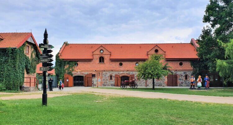 Widok na stajnie, budynek z czerwonej cegły i czerwoną dachówką, przed budynkiem stoją posągi ludzi, wokół spacerujący turyści - Skansen w Sierpcu